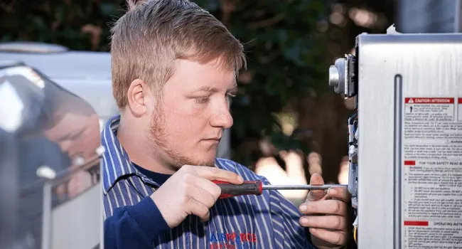Technician in blue striped shirt using screwdriver to repair electrical equipment outdoors in daylight.