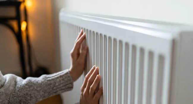 Person in gray sweater warming hands on a white radiator in a cozy indoor setting with soft lighting.