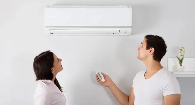 Man and woman controlling a wall-mounted air conditioner with a remote in a bright room.