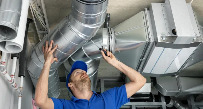 Technician in blue uniform inspecting and repairing metal HVAC ductwork with a screwdriver in a commercial building.