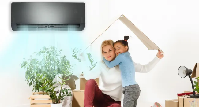 Mother and daughter sitting under cardboard roof near air conditioner cooling the room with green plants and boxes