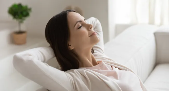 Woman relaxing peacefully on a white sofa with eyes closed and hands behind head in a bright living room.