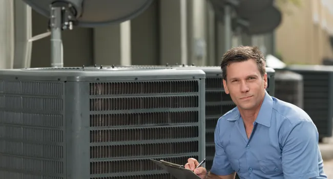 Technician inspecting HVAC unit outdoors with clipboard, wearing blue uniform and kneeling beside equipment