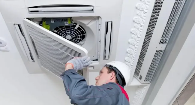 Technician in safety helmet and gloves installing or cleaning ceiling air conditioner filter in office space.