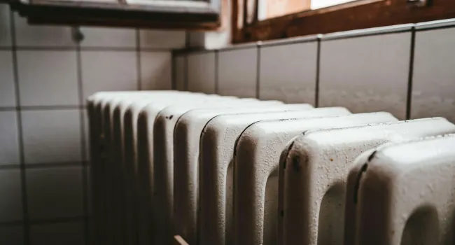 Close-up of an old white radiator against tiled wall near a wooden-framed window in a dim room