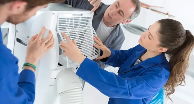 Three technicians in blue uniforms assembling or repairing a white air conditioning unit indoors.