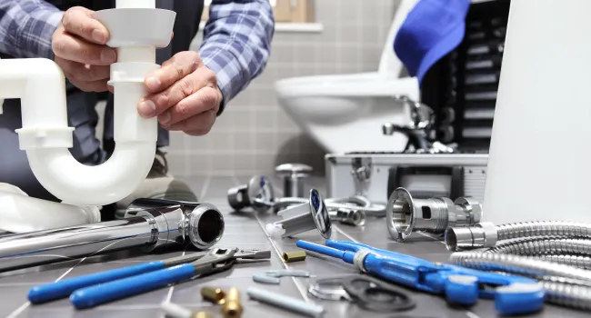 Plumber fitting a white PVC pipe under a bathroom sink with various plumbing tools scattered on the floor.