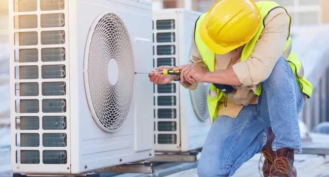 Technician in yellow hard hat and safety vest repairing outdoor air conditioning unit with screwdriver.