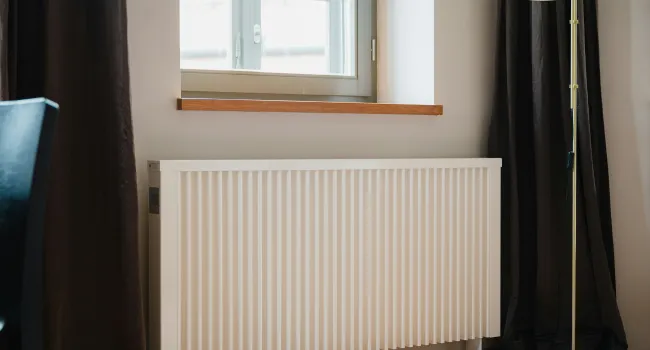 White vertical panel radiator below a window between dark curtains and a standing lamp in a room.
