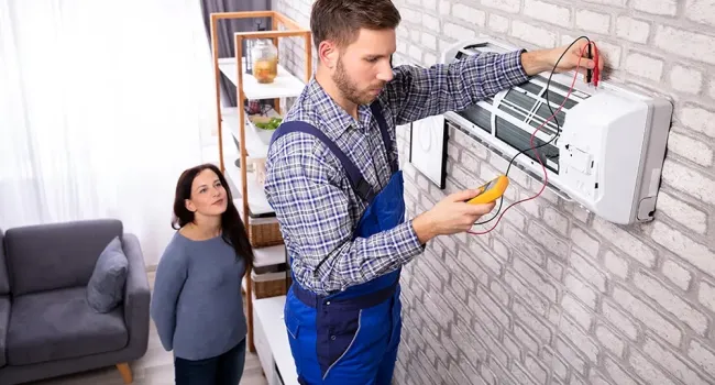 Technician in blue overalls repairing wall-mounted air conditioner while woman watches in living room.
