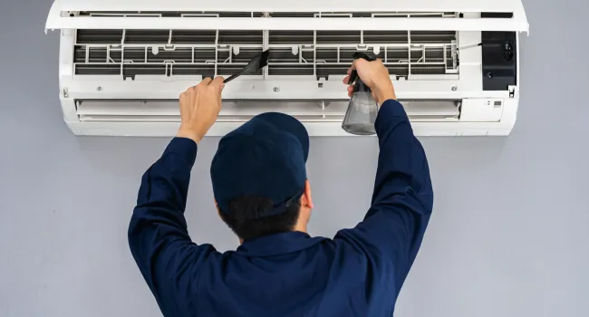Technician cleaning and maintaining a wall-mounted air conditioner using a brush and spray bottle