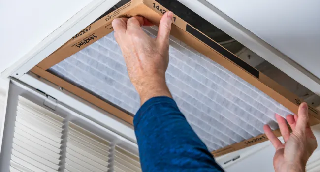 Person installing a rectangular air filter into a ceiling HVAC vent with white frame and grilles.