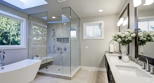 Modern bathroom with freestanding tub, glass shower enclosure, skylight, and double vanity with large mirrors and flowers.