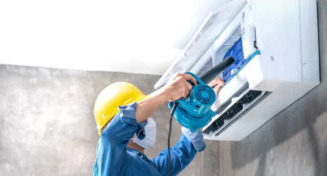 Technician wearing yellow helmet cleaning an indoor air conditioner with a blue blower device.