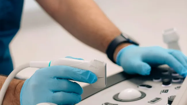Medical professional wearing blue gloves operating an ultrasound machine with control panel and probes.