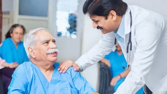 Doctor smiling and comforting elderly male patient in hospital setting with medical staff in background