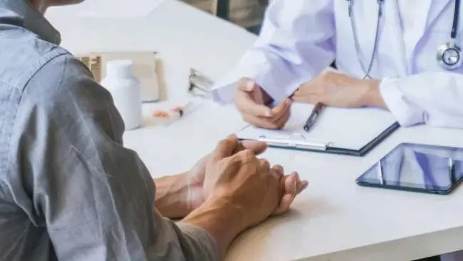 Patient consulting with doctor at desk with medical clipboard and tablet during health appointment.
