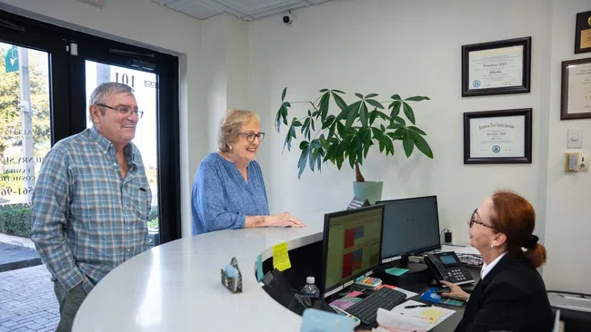 Senior couple smiles while speaking with receptionist at a clinic's front desk with certificates on the wall