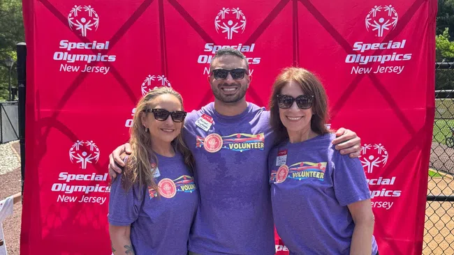 Three smiling volunteers wearing purple shirts with rainbow volunteer badges stand before a Special Olympics New Jersey backdrop.