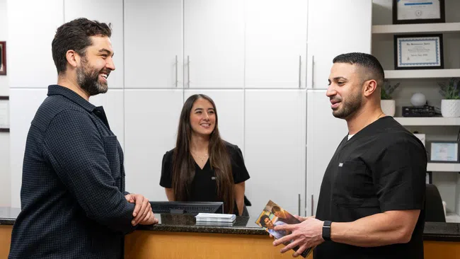 Two men speaking at a dental clinic reception desk with a female staff member smiling behind the counter