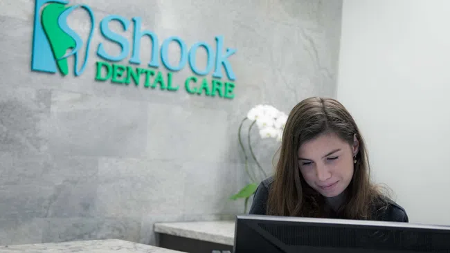 Receptionist working at Shook Dental Care front desk with logo and flower decoration in background