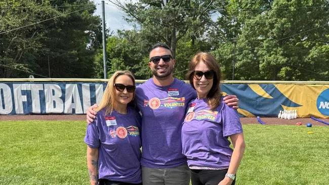Three volunteers in purple shirts standing on grass at a softball field with trees and blue sky behind them.