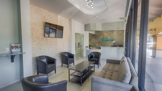 Modern waiting room with gray sofa, black chairs, coffee table, TV, and reception desk in a dental office.