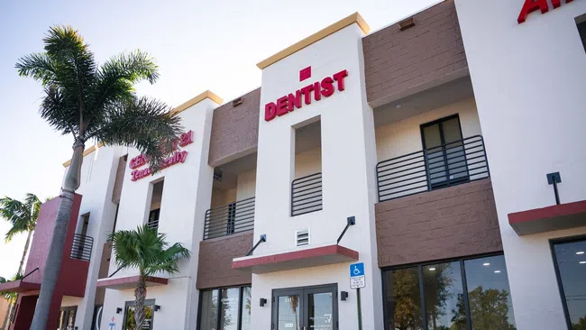 Modern dental clinic building exterior with palm trees and clear sky, featuring prominent dentist signage.