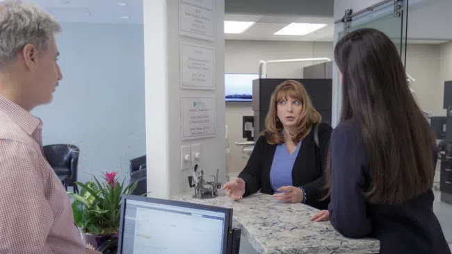 Two women speaking to a male receptionist at a healthcare or office reception desk in a professional setting.