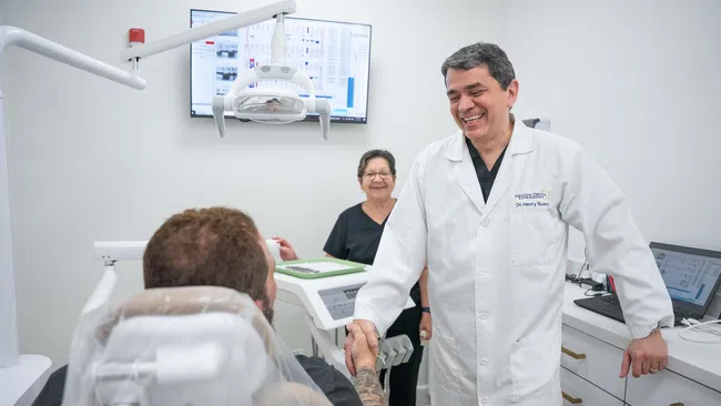 Dentist in white coat shaking hands with patient in dental clinic with assistant smiling in background