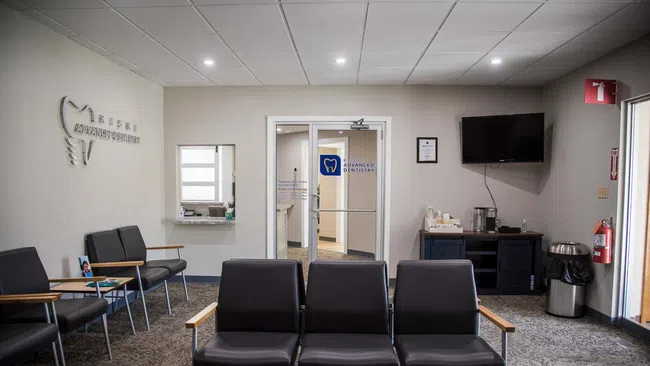 Modern dental office waiting room with black chairs, TV, water dispenser, fire extinguisher, and glass doors.