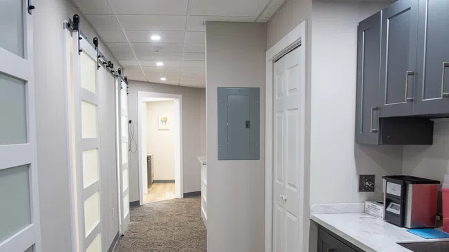 Modern hallway with carpeted floor, white sliding doors, gray cabinets, and bright ceiling lights in an office or clinic.
