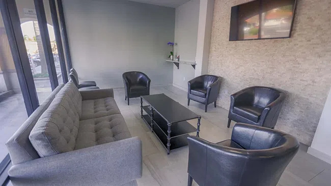 Modern waiting room with gray sofa, black leather chairs, glass windows, and a wall-mounted TV.