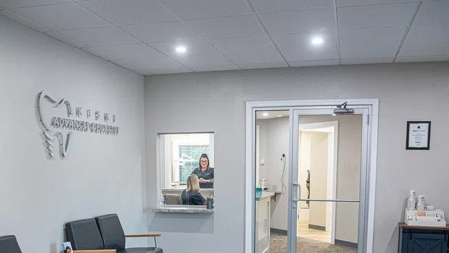 Modern dental office waiting room with black chairs, gray carpet, and reception windows with staff inside.