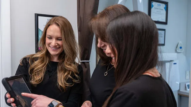 Three women smiling and interacting while looking at a tablet in an indoor setting with framed certificates.