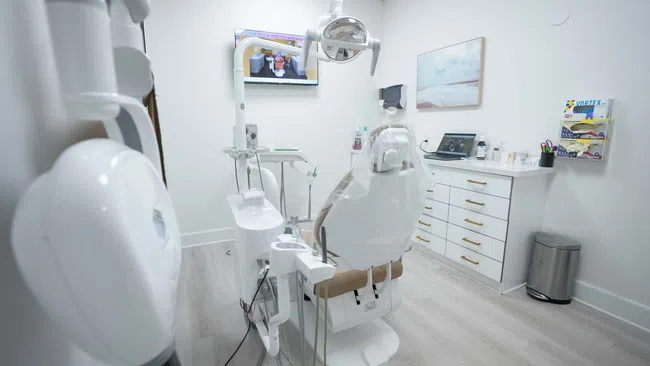 Modern dental clinic room with dental chair, equipment, TV screen, and clean white interior.