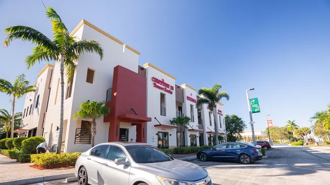 Modern commercial building with palm trees and parked cars under a clear blue sky on a sunny day