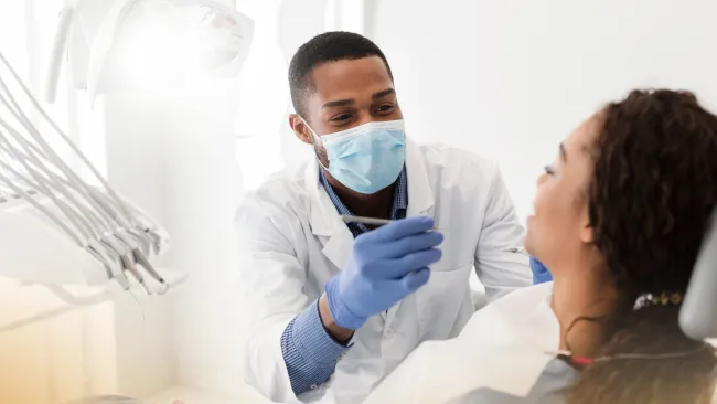 Male dentist wearing mask and gloves examining female patient's teeth in bright dental office.