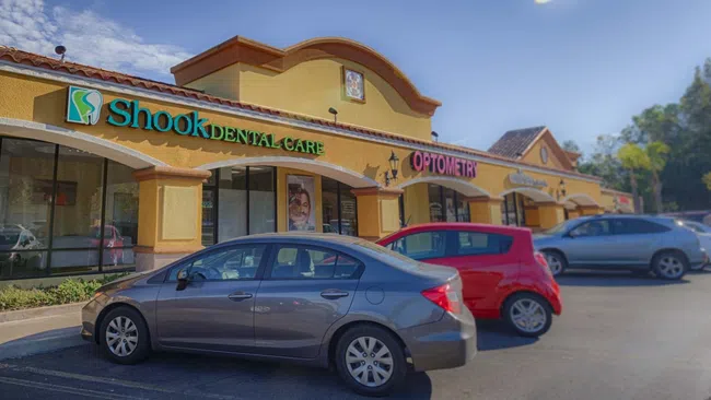 Shopping plaza with Shook Dental Care and Optometry signs, several parked cars, and blue sky.