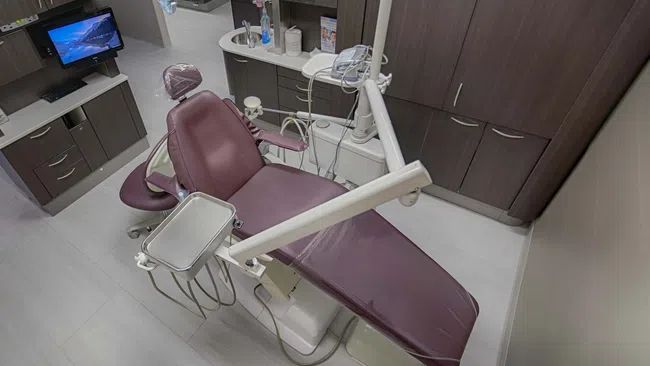 Modern dental clinic room with maroon dental chair, equipment, computer monitor, and wood cabinetry.