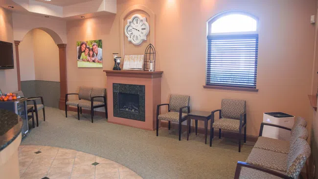 Cozy waiting room with chairs, a fireplace, wall clock, window, and neutral decor in soft lighting.