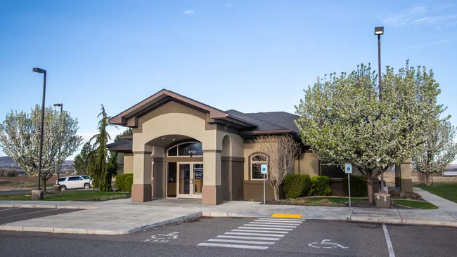Modern beige single-story office building with arched entrance surrounded by blooming trees and parking spaces.