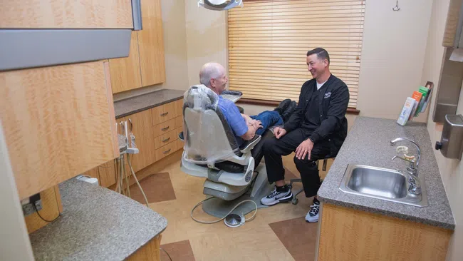 Dentist talking and smiling with elderly male patient seated in dental chair in modern clinic room.