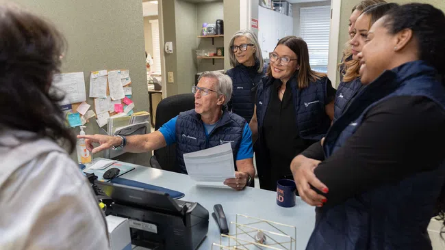 Team of six coworkers in blue vests collaborating in an office around a desk with papers and computer.