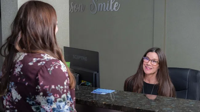 Receptionist smiling and assisting a woman at a clinic front desk with computer and sign behind.
