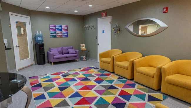 Colorful waiting room with yellow chairs, purple loveseat, geometric rug, water cooler, and decorative wall mirror.