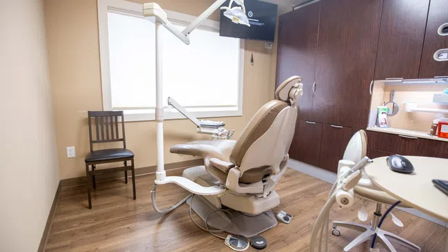 Modern dental office room with beige patient chair, equipment, wood cabinets, and hardwood flooring under bright light.