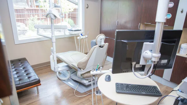Modern dental clinic room with reclining chair, dental tools, computer, and window with outside view.