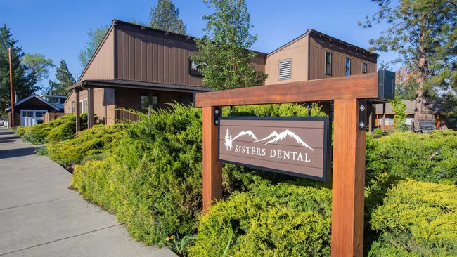 Sisters Dental sign with mountain logo outside brown building surrounded by green bushes and trees on sunny day