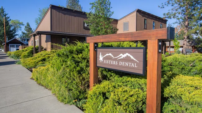 Modern brown Sisters Dental office building with wooden sign and green bushes under clear blue sky.